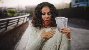 Bir şehir caddesinde Filipinli peso banknotları tutan kadın, bir süveter giyerken duygularını ifade ediyor, bulanık şehir geçmişi ve parmaklıklarla çevrili..