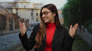 Kırmızı bluzlu ve siyah ceketli İspanyol kadın, şehir arka planında elleri açık bir ortamda iyimser bir şekilde gülümsüyor..