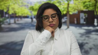 Kadın şehir caddesinde güneş ışığı ağaçların arasından süzülüyor, gözlük ve beyaz bluz giyiyor, şehir ortamında çeşitliliği ve modayı kucaklıyor..