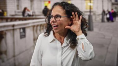 Gözlüklü ve kıvırcık saçlı bir kadın şehir sokaklarında durup dinlerken, şehir seslerinden etkilenmiş, canlı enerji tasvir eden ve dışarıdaki iletişimi meşgul eden etkileyici bir yüz..