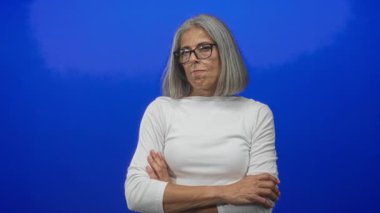 Gri saçlı, gözlüklü ve beyaz parmaklı bir kadın mavi stüdyoda barış işareti yapıyor..