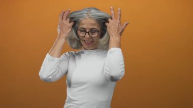 Turuncu stüdyo stüdyosunda, gri saçlara ve boyuna yakın ellere dokunan kadın tebessümle gülümsüyor beyaz bluzuyla; refah memnuniyeti.