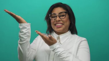 Gözlüklü ve beyaz gömlekli genç İspanyol kadın parlak bir şekilde gülümsüyor. Parlak yeşil bir arkaplan üzerinde ellerini uzatıyor. Olumlu ve coşkulu bir hava yayıyor..