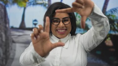 Açık şehir ortamında gülümseyen bir kadın, bir resmi çerçevelemek için ellerini kullanıyor, canlı bir sokak arka planında pozitifliğini ve yaratıcılığını ifade ediyor..