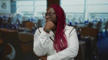 Pembe örgülü bir kadın restoran ortamında düşünceli bir şekilde poz veriyor. Gözlük takıyor ve şık bir bilezik takıyor. Modern ve rahat bir iç mekan yaratıyor..