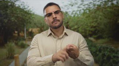 Gözlüklü ve bej gömlekli bir adam, elini inceleyen ve bir orman ortamında dikilirken parmağıyla avuç içini gösteren;.