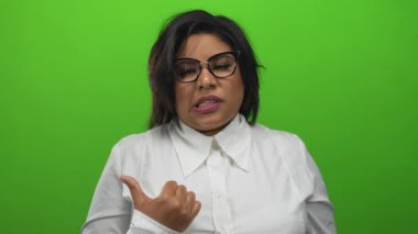 Canlı yeşil arka plana karşı baş parmağıyla hareket eden gözlüklü ve beyaz gömlekli bir kadın yüz ifadesiyle şaşkınlığı gösteriyor.