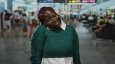 Havaalanı terminalinde oturan yeşil kazaklı kadın, mutlu bir ifadeyle, kalabalık gezginlerle çevrili, kapalı bir havaalanı ortamında..