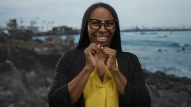 Sahilde duran, sarı tişörtlü ve gözlüklü bir kadın açık hava ortamında kayalık bir sahil ve okyanus manzaralı dramatik bir ifade takınıyor..