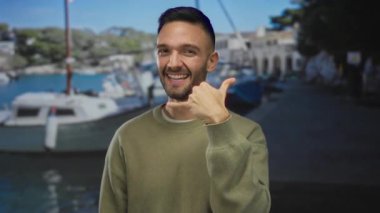 Genç adam telefon ediyor ve gülümseyerek deniz ve teknelerin yakınındaki gezinti alanında işaret ediyor.