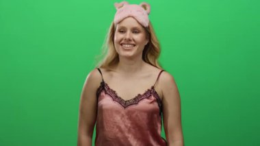 Gecelikli bir kadın canlı yeşil bir arka plana karşı gizli bir jest yapıyor pembe bir uyku maskesi takıp neşeyle gülümserken neşeli bir gizlilik ve entrika sergiliyor..