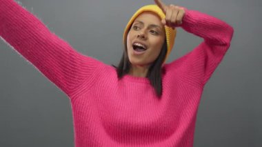 Pembe kazak giyen ve sarı şapkalı bir kadın gri bir arka plana sevinçle bakıyor.