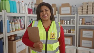 Yansıtıcı yelek giyen genç bir kadın bağış merkezinde pano tutuyor. Kutular ve ürünlerle çevrili. Destek veren bir ortamda hayır işlerine olan bağlılığını gösteriyor..