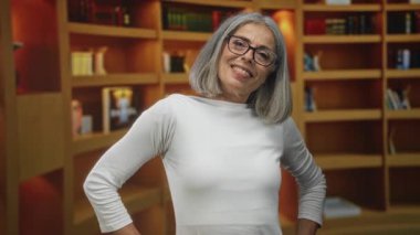 Orta yaşlı, gri saçlı bir kadın, kütüphane binasında kitapların arasında sırıtarak gülümsüyor. Gözlük ve beyaz bir bluz giyiyor..