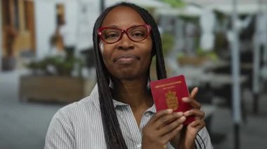 Elinde Danimarka pasaportu olan bir kadın teras kafeteryasında güvenli bir şekilde gülümsüyor şehir sokaklarının bulanık arka planıyla..