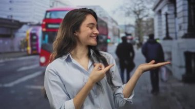 Kadın gülümsüyor ve arkasında kırmızı çift katlı otobüsle şehir caddesinde avucunun içini işaret ediyor; iyimserlik.