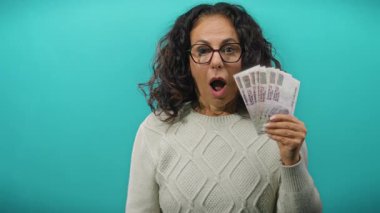 Orta yaşlı, gözlüklü, elinde Mısır banknotları olan mavi arka planda sürpriz bir ifadeyle para hevesi ve kültürel bağlam gösteren bir kadın..