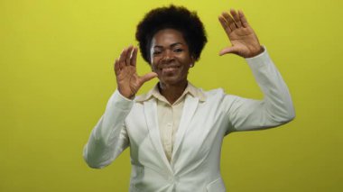 Sarı arka planda beyaz ceketle gülümseyen bir kadın stüdyo ortamında elini kaldırarak sevinçten havalara uçuyor..