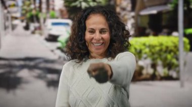 Kıvırcık saçlı, kazak giymiş, güneşli bir caddede gülümseyen bir kadın canlı bir şehir gününde pozitiflik ve güven saçıyor..