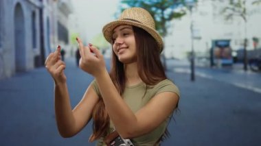 Açık bir şehir sokağında iki eliyle kaba bir jest yapan bir kadın neşeli havasını şapkası ve kamerasıyla sergiliyor..