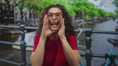 Kanal köprüsünde elleri ağızdan ağıza kırmızı gömlek ve gözlük takan bir kadın. İletişim heyecanı..