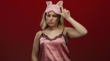 Pembe gece elbisesi ve göz maskesi takan kadın canlı kırmızı bir arka plana karşı poz veriyor, neşeli bir ifadeyle ve rahat bir tavırla yatma modasını sergiliyor..