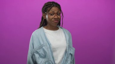 Gözlüklü, gülümseyen ve kameraya yaslanan bir kadın stüdyoda mor arka plan ve kot gömlekli, kendine güvenen..