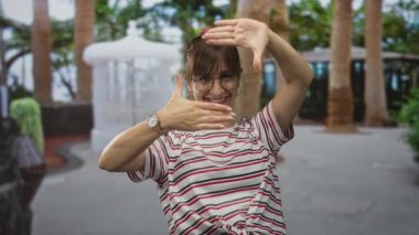 Elleri dikdörtgen şeklinde çerçeveleyen ve palmiye ağaçlarıyla dolu bir parkta fıskiyenin yanında gülümseyen, gözlük takan ve çizgili gömlek giyen, yaratıcı neşe veren bir kadın..