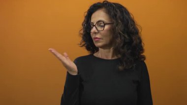 Orta yaşlı kıvırcık saçlı ve gözlüklü İspanyol kadın, bir stüdyoda avucunu ileri doğru açık tutuyor; reddediyor..