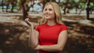 Sarışın genç bir kadın, yeşil ağaçlarla çevrili güneşli bir parkta, kırmızı gömleği ve kotuyla güvenle gülümsüyor..