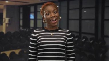 Kadın, spor salonunun içinde halter askısı ve spor aletleriyle çizgili süveter giyerken gülümsüyor; onaylanmıyor.