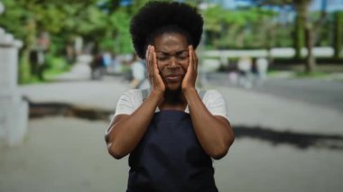 Açık şehir caddesinde elleri açık önlük giyen bir kadın, bulanık arka plan ve yeşil yeşillikler arasında duygu ve içgözlem taşıyor..