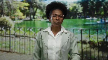 Parkta gözlüklü bir kadın, takım elbise giymiş, göllü ve yemyeşil ağaçlı sakin bir yaz ortamı yaratıyor..