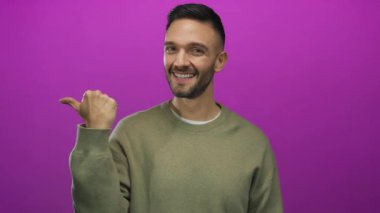 İspanyol genç adam gülümsüyor ve baş parmağını parlak pembe bir arka plana doğrultuyor, günlük bir süveter giyiyor, stüdyoda pozitiflik yayıyor..