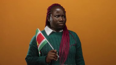 Woman with suriname flag over orange background makes thoughtful gesture implying realization or idea while wearing a green sweater and pink braids.