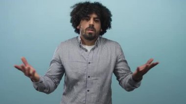 Kıvırcık saçlı ve sakallı bir adam açık mavi bir stüdyoda açık avuçla omuz silkiyor. Çek gömlek giyiyor ve kurnazlık taslıyor..