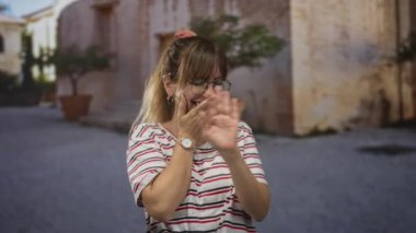 Gözlük ve çizgili tişört giyen, eski binanın önünde gülerken ağzı elle kapatan kadın, eğlence utangaçlığı..