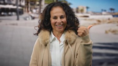 Kıvırcık saçlı sıradan bir kadın, deniz kenarında başparmağını sallayarak gülümserken güneşli sahilde güzel, olgun, İspanyol bir varlığı yakalıyor..