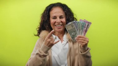 Orta yaşlı bir kadın elinde Hint rupisi notları tutarak parlak sarı bir zemin üzerinde kaya jestleri yaparak heyecan ve refahı ifade ediyor..