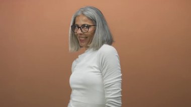 Parmaklarıyla işaret eden ve stüdyoda gözlük takan terracotta duvarı ve beyaz balıkçı yakalı, gri saçlı ve gülümseyen bir kadın..