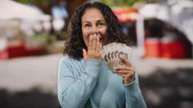 Şehir sokaklarında orta yaşlı bir kadın sürpriz ve neşe dolu bir ifadeyle Meksika pezosu tutuyor..