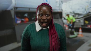 İnşaat alanında örgü ören kadın, yüksek görüş açısına sahip yelek ve ekipman giyen işçilerin yanında sürpriz bir ifade sergiliyor..