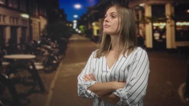 Gece sokak lambalarının altında kolları çaprazlanmış beyaz çizgili elbiseli kadın bisikletlerin yanında..