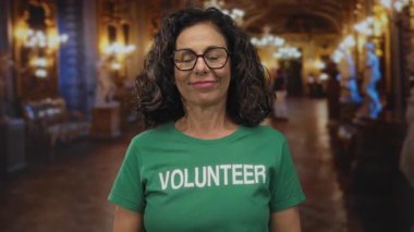 Müze salonunda orta yaşlı İspanyol kadın gönüllü yeşil tişört giyip dil uzatıyor, mizah nişanı yapıyor..