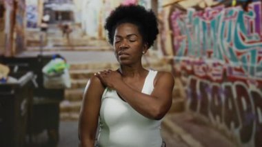 Kadın sokak ortasında graffiti ile geziyor, beyaz atlet giyiyor, düşünceli görünüyor, dayanıklılık ve direnç gösteriyor..