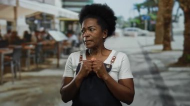 Önlüklü kadın hareketli bir açık hava restoranının terasında duruyor. Canlı bir şehir yemeği ve sokak hayatı sahnesi sergiliyor..