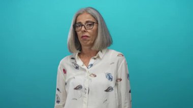 Gözlüklü ve desenli bluz giyen kadın, dil dışarı ve kollar hafifçe deniz mavisi stüdyosunda genişletilmiş; sessiz can sıkıntısı.