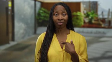 Sarı tişörtlü, kıvırcık saçlı bir kadın sokak arka planında açık hava manzarasını işaret ediyor..