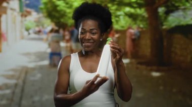 Elinde losyon şişesi olan bir kadın ağaçlarla bir caddede gülümsüyor ve arka planda insanlar güneşli bir günde beyaz bir bluz giyiyor..