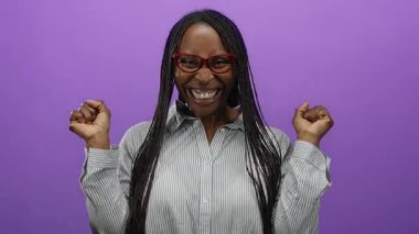 Kadın canlı mor bir arka plana karşı heyecanla kutluyor gözlük takıyor ve neşe ve pozitiflik taşıyan çizgili bir gömlek giyiyor..